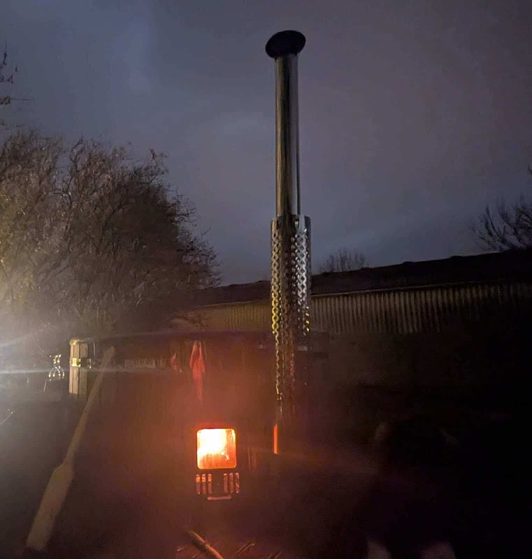 Nighttime outdoor scene showing glowing wood stove chimney with metal heat shield beside leafless trees and dark building silhouette
