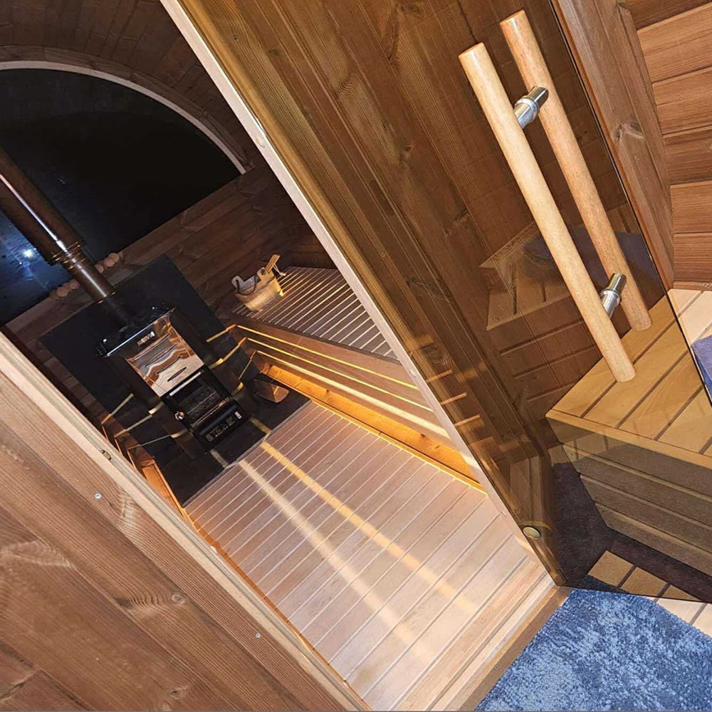 Interior view of a wooden sauna with built-in benches, a black wood stove, and glass door handles