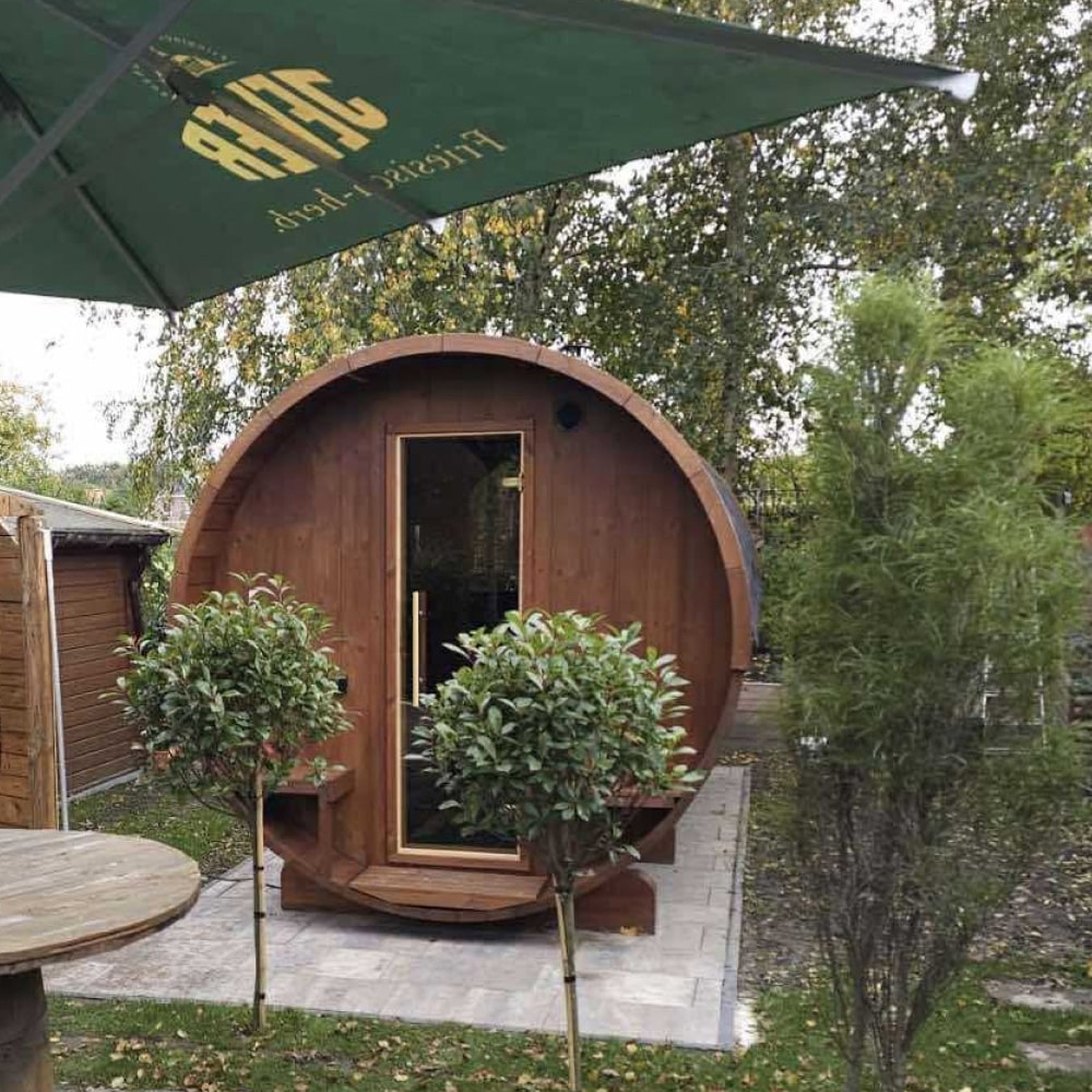 Outdoor view of small barrel-shaped wooden sauna with glass door and green umbrella in garden setting