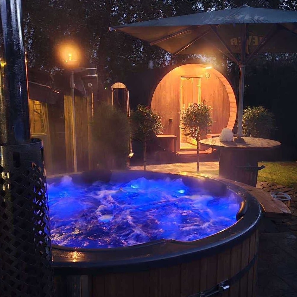 Nighttime view of a wooden hot tub with blue LED lights, steam rising, and a barrel sauna in the background