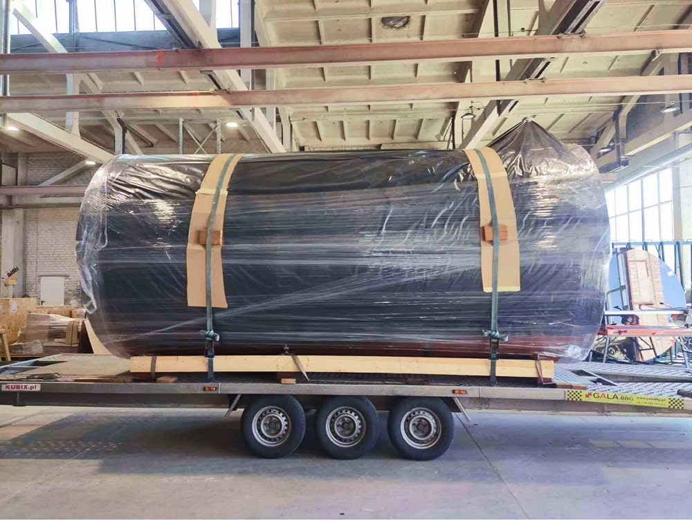 Large cylindrical object wrapped in plastic and secured with straps on flatbed trailer inside industrial warehouse
