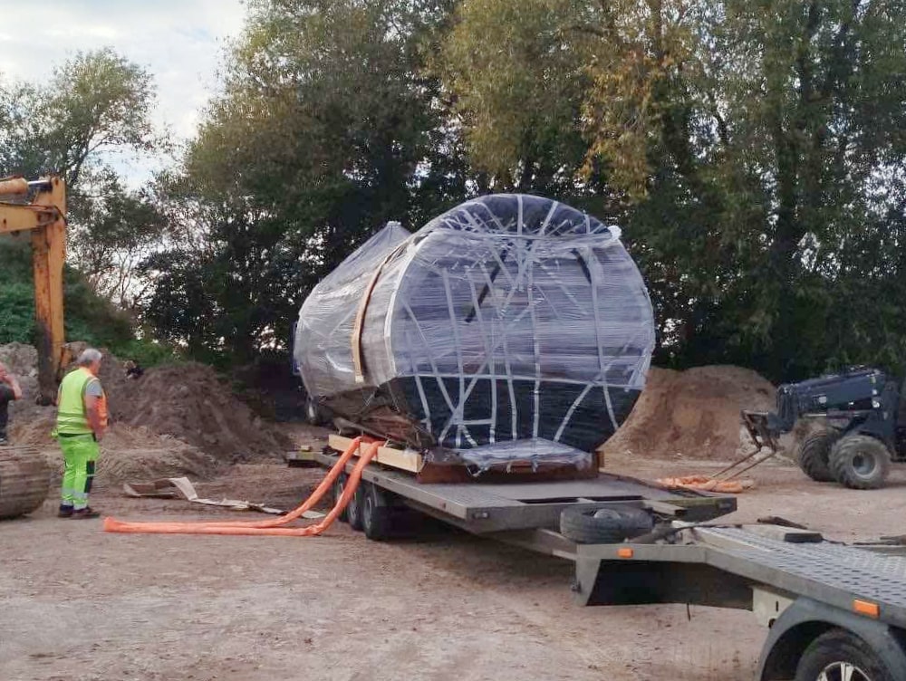 Outdoor view of large barrel sauna wrapped in plastic on trailer bed with workers and construction equipment nearby