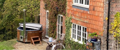 Round wood-fired hot tub with integrated stainless steel heater, installed beside a brick house with garden seating area.