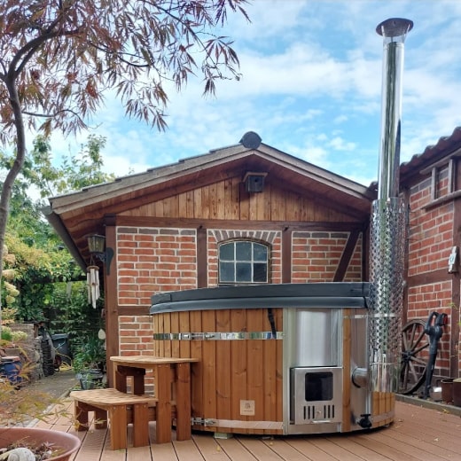 Outdoor view of a round wood fired hot tub with stainless steel stove and wooden steps on wooden deck by brick building