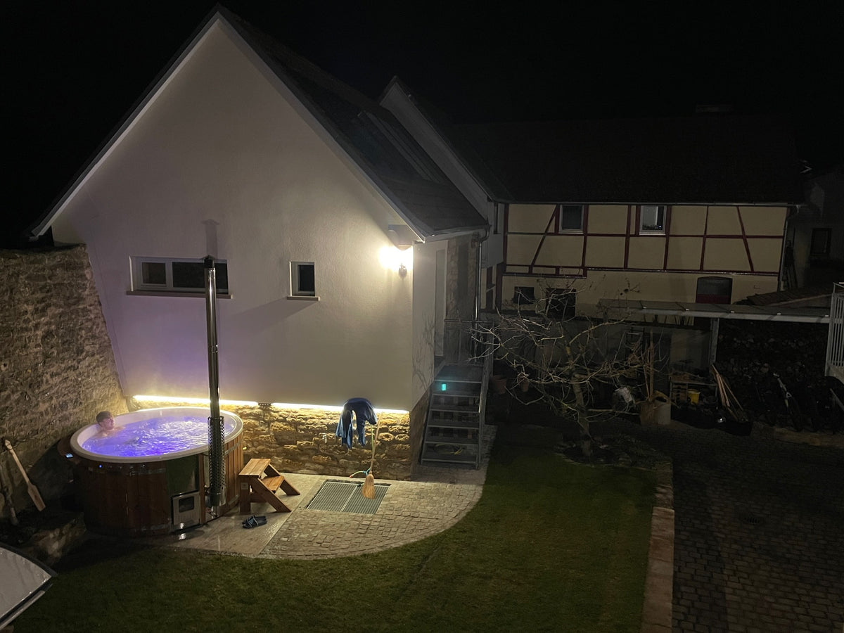 Naktinis vaizdas į apšviestą lauko kubilą šalia akmens ir tinkuoto namo su gretimu sodu ir mediniais laiptais