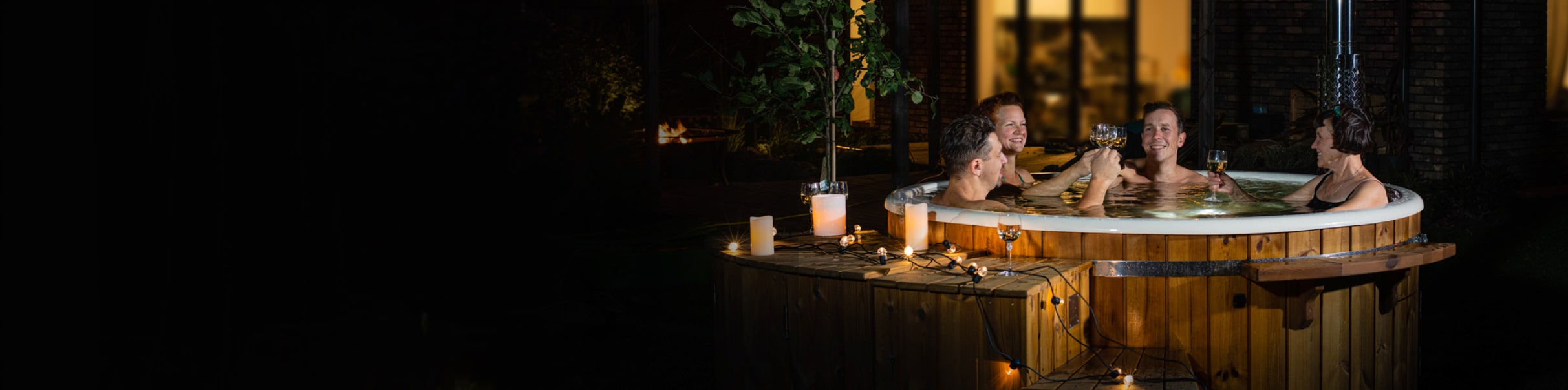 Night view of four people relaxing in a wood-fired hot tub with warm lighting and wooden exterior panels