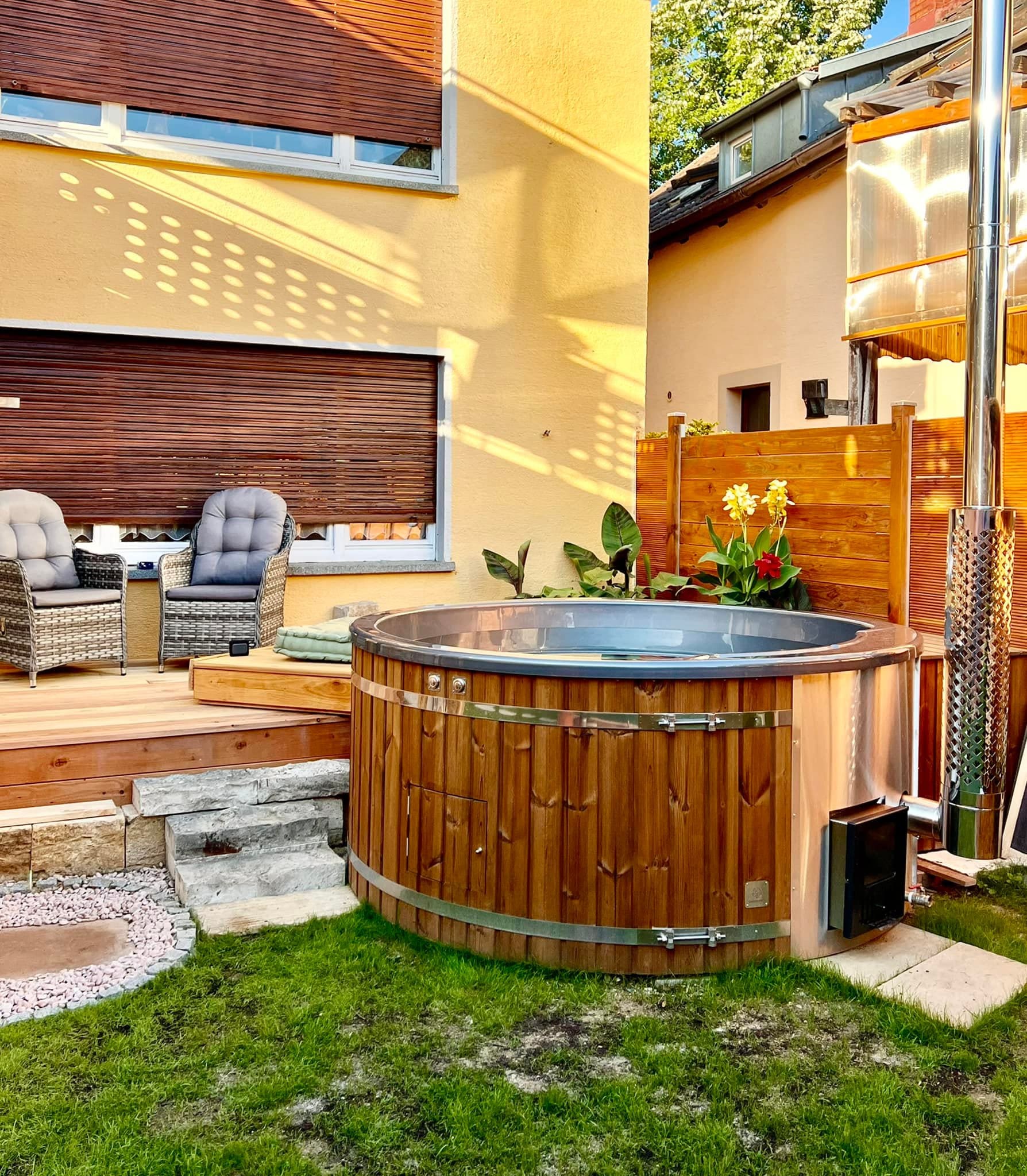 Gardenvity wood fired hot tub in urban garden beside yellow house with patio furniture.