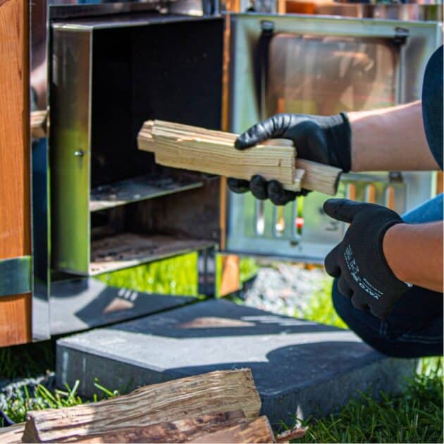 Person loading firewood into a stainless steel wood-fired hot tub stove outdoors.