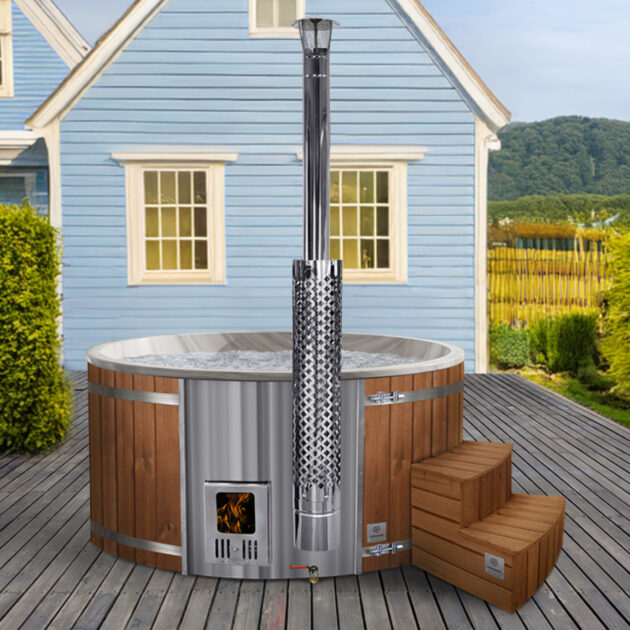 Round wood-fired hot tub with stainless steel stove and wooden steps on a terrace in front of a blue house.
