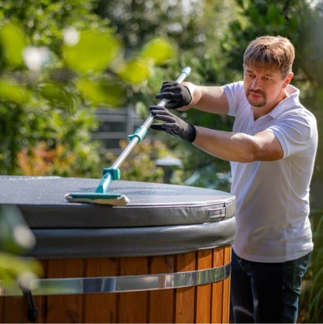 Man cleaning an insulated hot tub cover using a brush in an outdoor garden setting