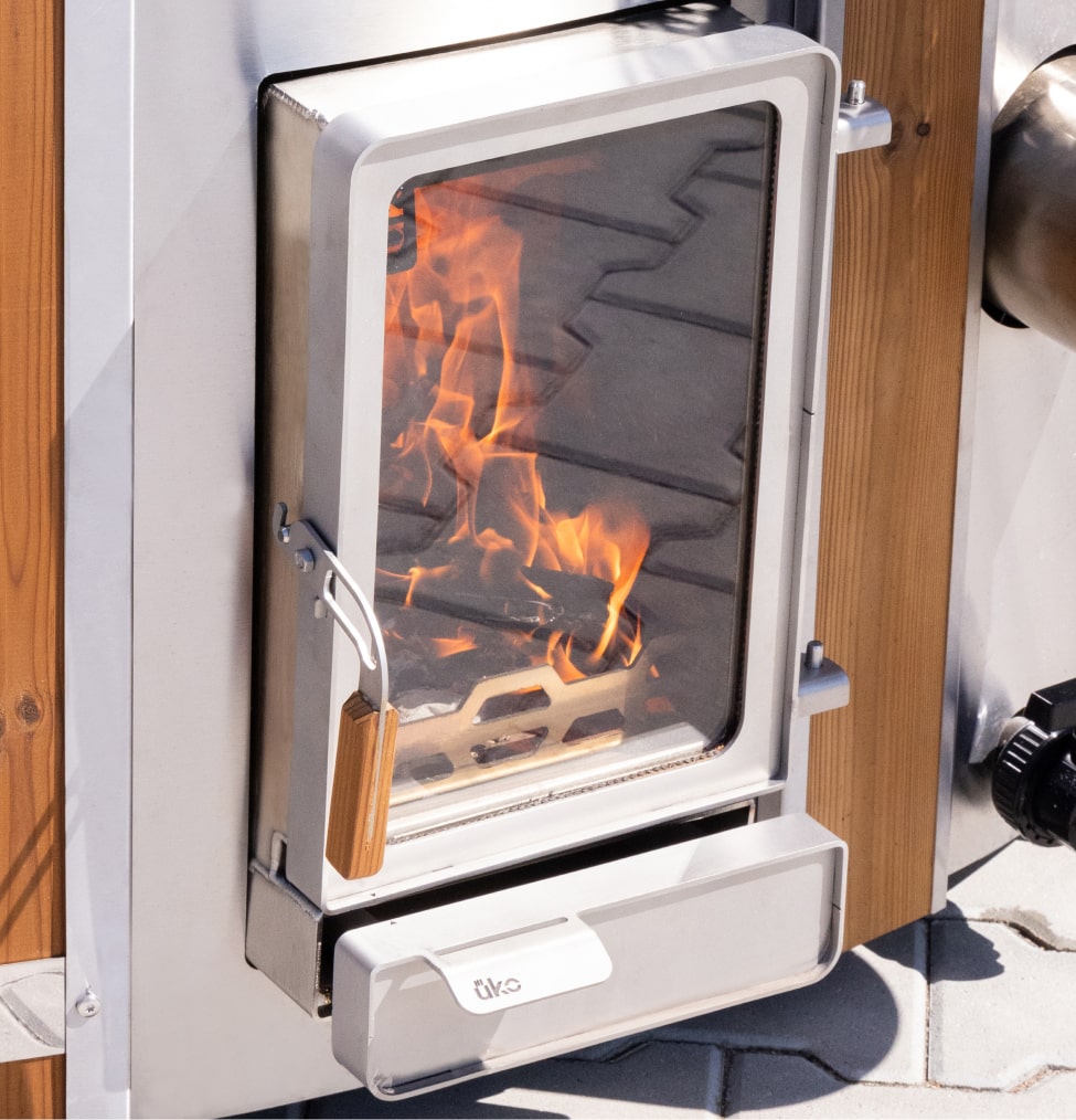 Close-up of stainless steel wood-fired hot tub heater door with glass window and visible flames inside.