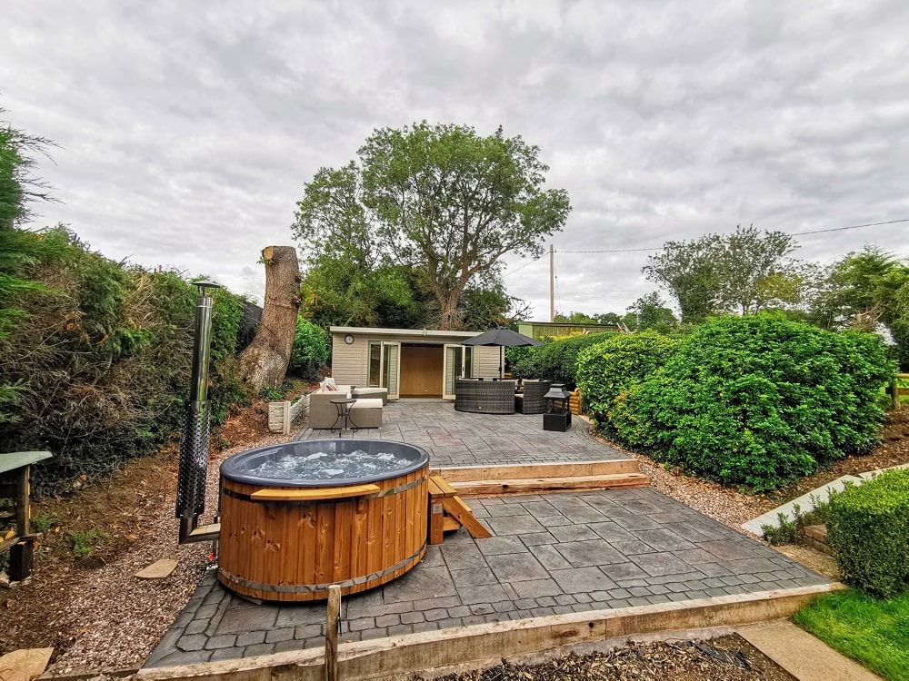 Gardenvity wood fired hot tub in patio area with rustic outdoor furniture and stone flooring.