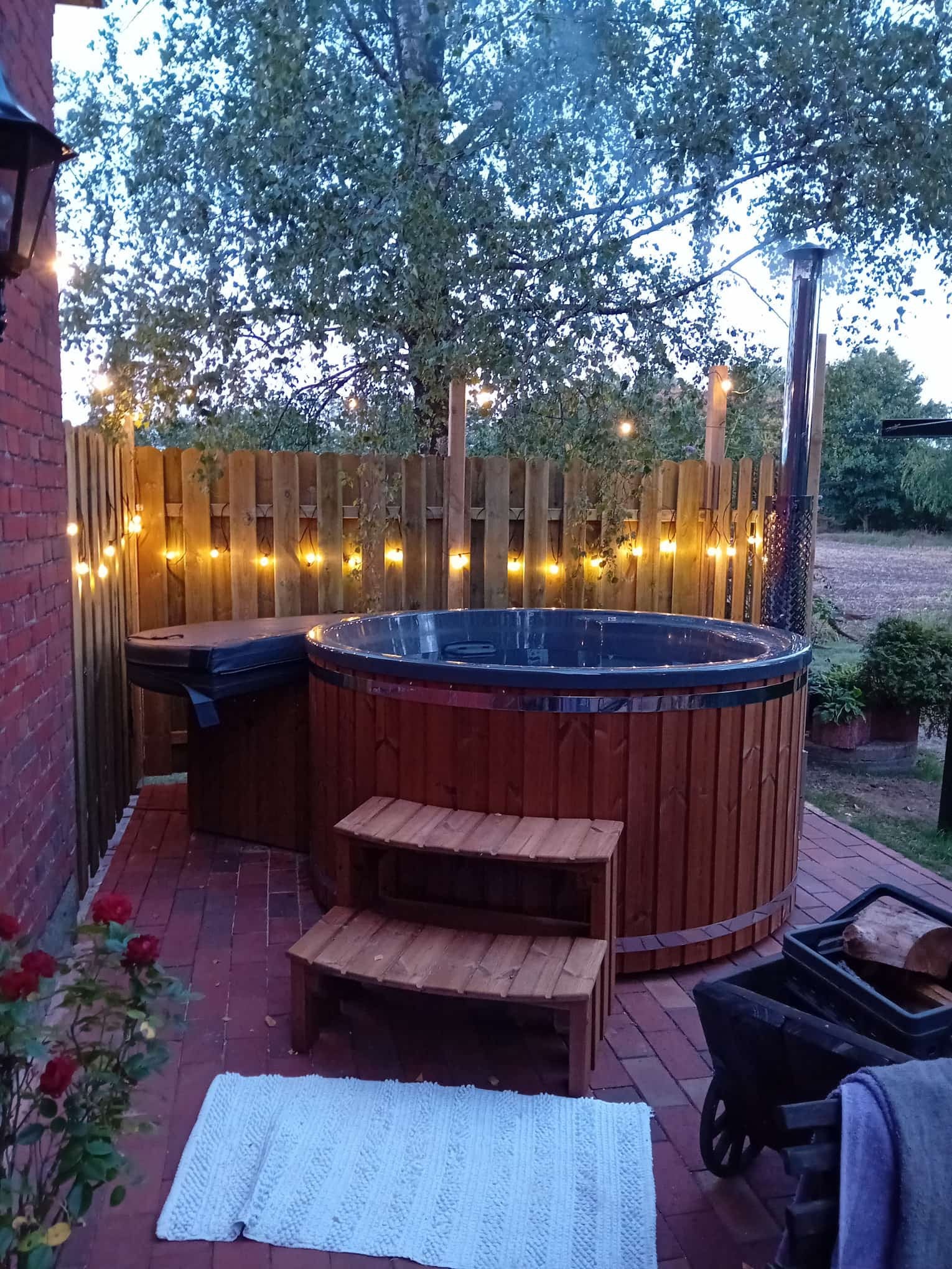 Gardenvity wood fired hot tub surrounded by fairy lights on wooden deck at night.