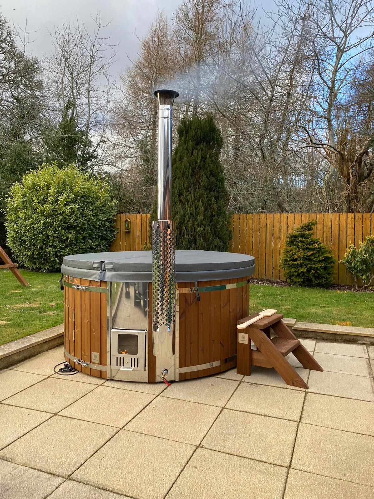 Gardenvity wood fired hot tub on tiled terrace surrounded by wooden fence and garden plants.