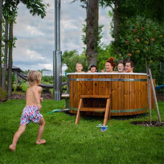 Family enjoying a round wood-fired hot tub outdoors, with children playing nearby in a garden setting.
