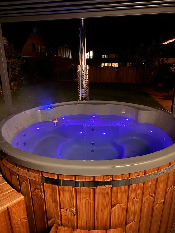 Wood-fired hot tub illuminated with blue LED lighting at night, highlighting underwater and exterior lights.