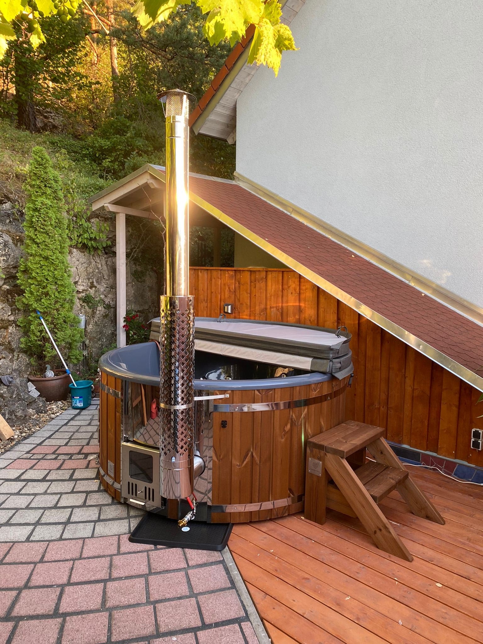 Gardenvity wood fired hot tub beside white house, stainless steel stove and mountain backdrop.