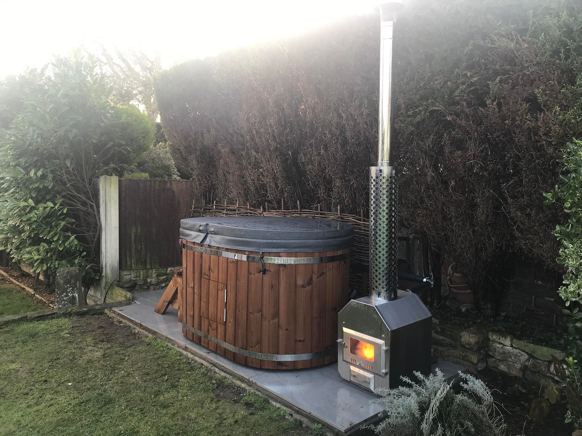 Gardenvity wood fired hot tub installed in private backyard with hedge and classic wooden design.