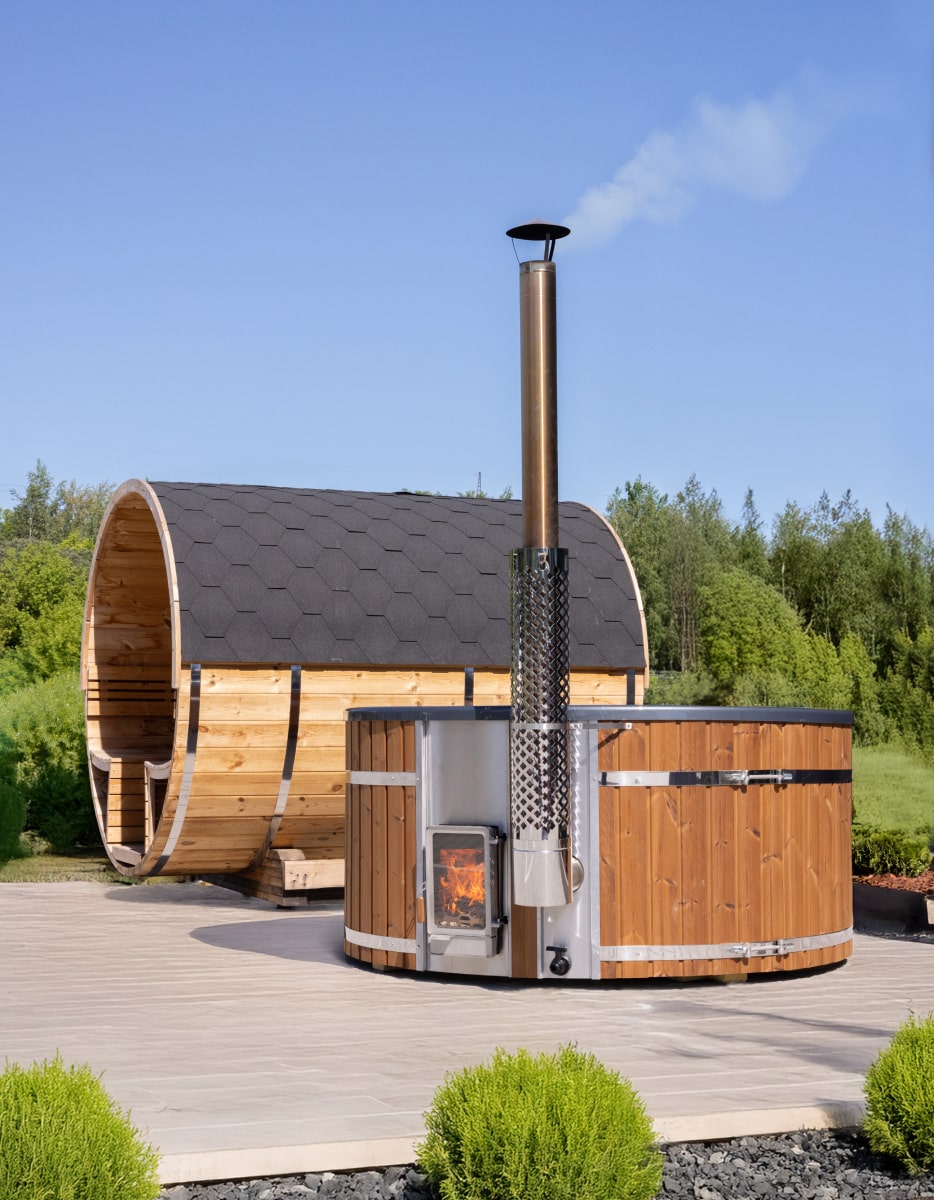 Round wood-fired hot tub with integrated stainless steel heater and chimney, placed beside a barrel sauna outdoors.