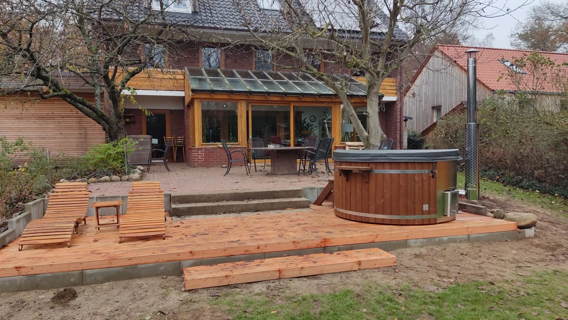 Gardenvity wood fired hot tub on wooden decking in a rustic backyard patio with brick house