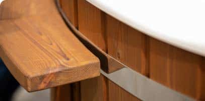 Close-up of a wooden bench attached to the side of a round wooden hot tub with a metal band and a white rim on top.